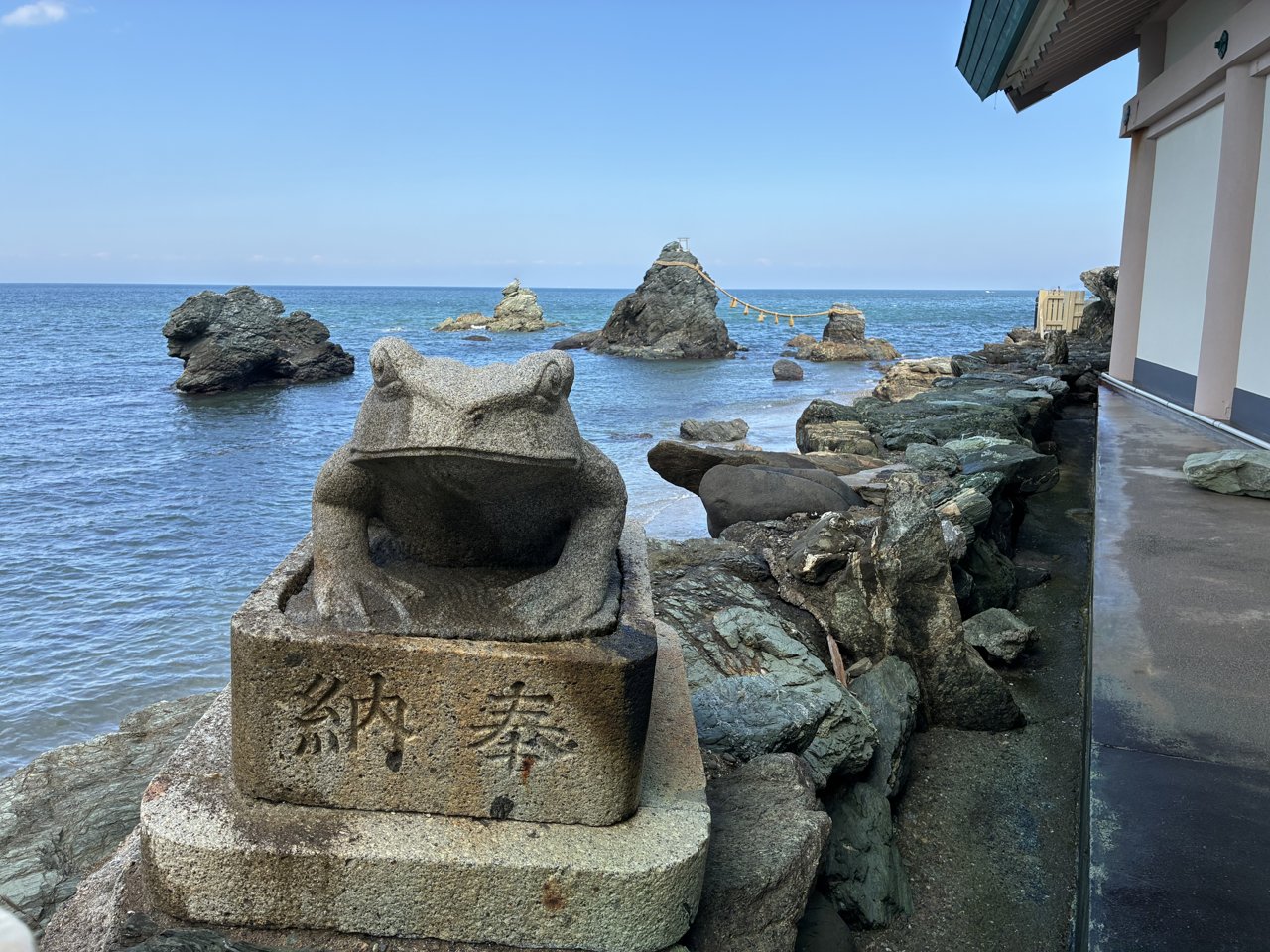奉納と彫られた台に座る二見興玉神社の石のカエルと、奥の夫婦岩